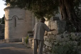 An elderly villager traces a faded carving on a stone wall near Clarensac’s church, morning light highlighting the textured stones and wild herbs nearby.