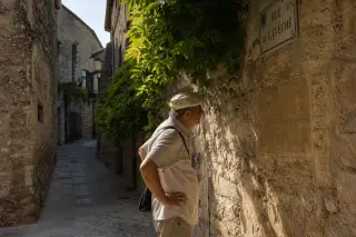 Elderly person reading an inscription on an old stone wall in a sunlit, narrow village alley lined with vines.