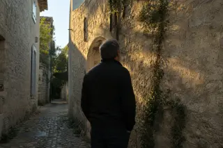 Man observes weathered stone wall with old inscription in narrow Clarensac alley at dusk, lit by soft Mediterranean evening light