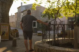 Middle-aged person from behind touching an old village fountain, warm golden light, limestone walls, crates, and festival signs around a southern French square.