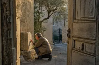 Narrow village lane bathed in golden evening light, seen from behind a half-open wooden door; an older local examines a weathered stone, framed by olive branches and rustic stone walls.
