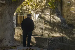 Mature local standing by an old limestone fountain in a Gard village at sunrise, viewed from behind, with sunlight illuminating stone textures and autumn leaves.
